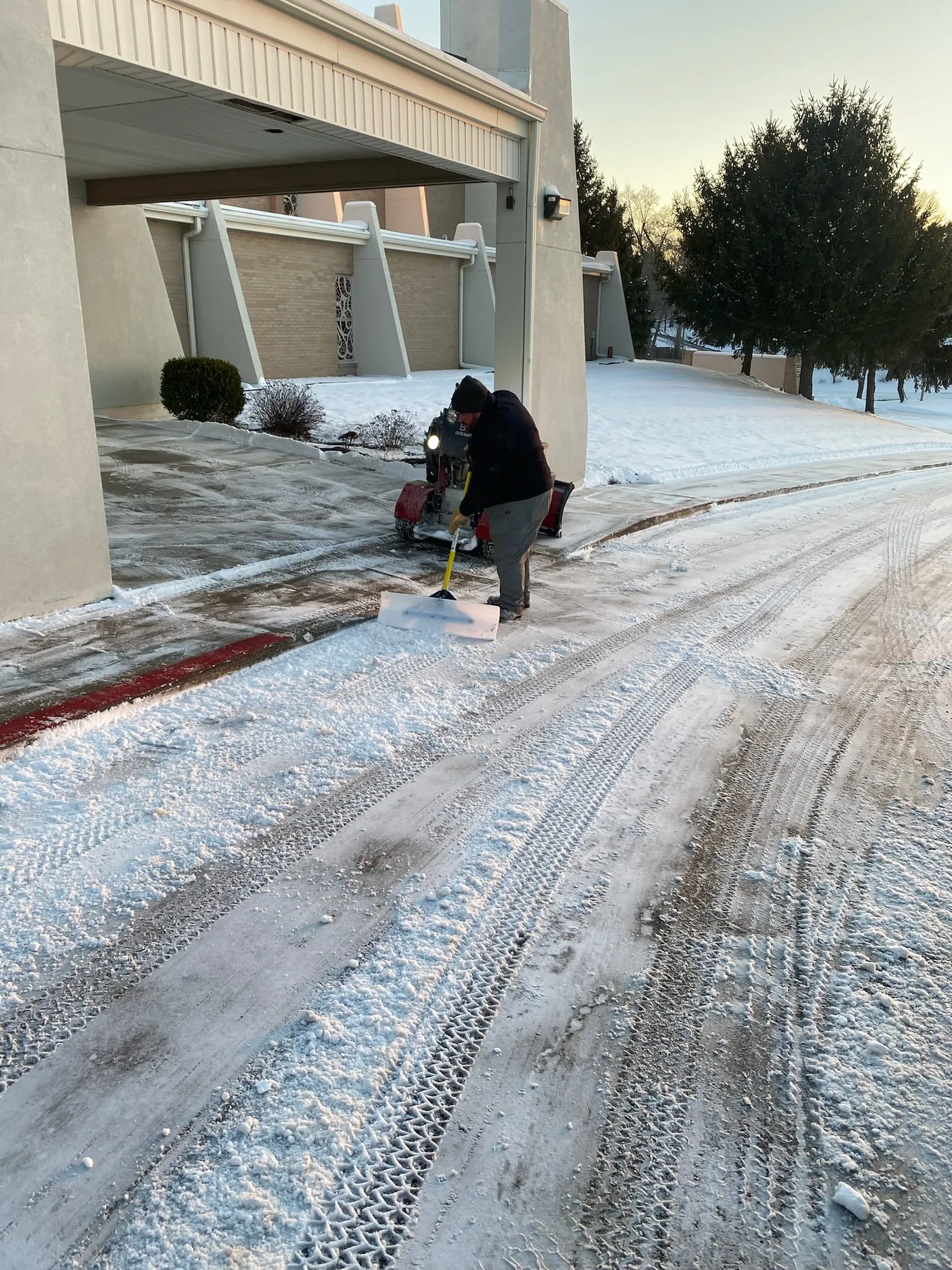 Crew member shoveling a walkway with a tracked snow blower at dawn